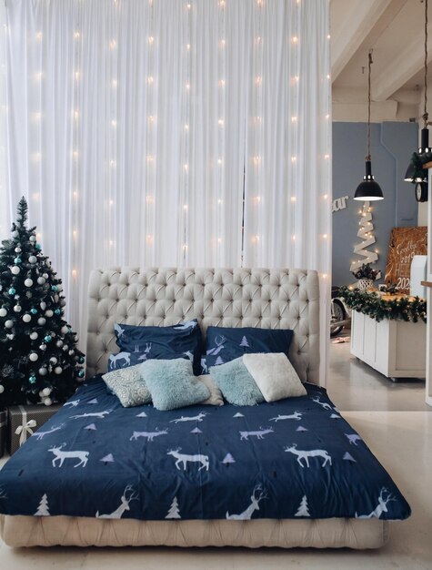 View over made bed with beautiful bedsheet with deer and cushions. The wall decorated with garland. The bedroom decorated for Christmas with fir tree.