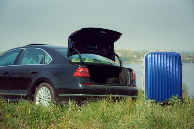 View of modern black car and blue suitcase on the river's side in sunny day.