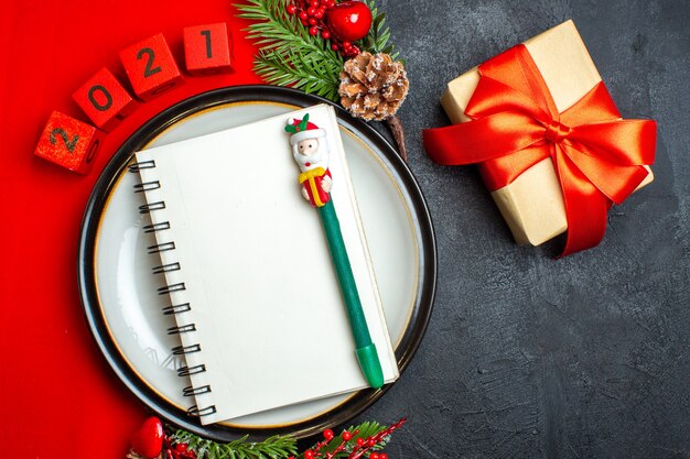 Above view of New year background with spiral notebook on dinner plate decoration accessories fir branches and numbers on a red napkin and gift with red ribbon on a black table