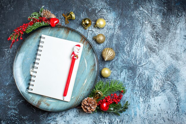 Above view of notebook with pen on blue plate decorative accessories on the right side on dark background