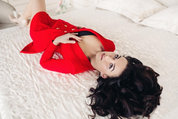 Вид красивой и энергичной женщины в красном платье с низким вырезом, лежащей на спине и отдыхающей на белой кровати, смотрящей в камеру сексуальная брюнетка с волнистыми волосами, идеальный макияж концепция моды и страсти