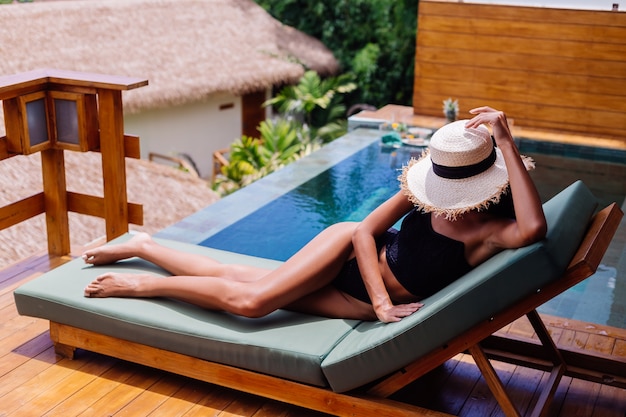 Free photo view of pretty tanned woman in black vintage bikini lies on green sun bed at amazing villa at sunny day, resting, enjoying vacation