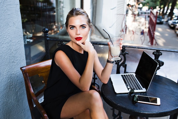 Above view pretty woman in black short dress sitting at table on terrace in cafeteria. She is looking to camera.