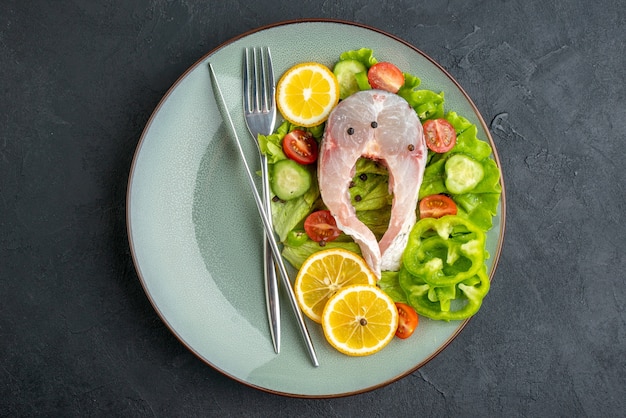 Above view of raw fish and fresh vegetables lemon slices and cutlery set on a gray plate on black surface with free space