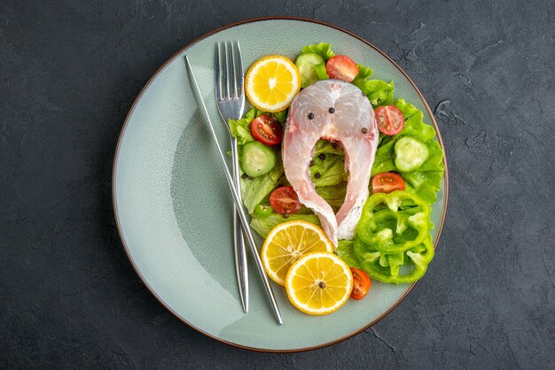 Выше вид сырой рыбы и свежих овощей, ломтики лимона и столовые приборы, установленные на серой тарелке на черной поверхности со свободным пространством