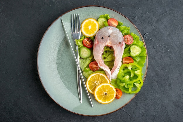 生の魚と新鮮な野菜のレモンのスライスとカトラリーが、空きスペースのある黒い表面の灰色の皿に置かれたビューの上