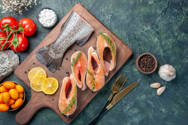 Above view of raw fishes lemon slices greens pepper on wooden cutting board tomatoes cutlery set on dark table