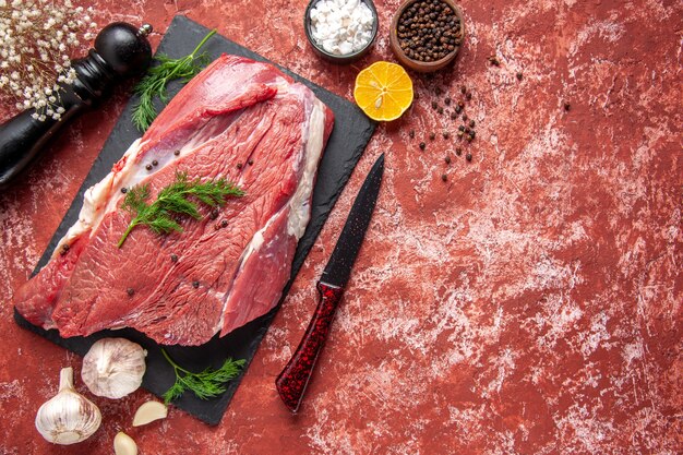 Above view of raw fresh red meat with green and pepper on black board knife wooden hammer salt lemon on oil pastel red background