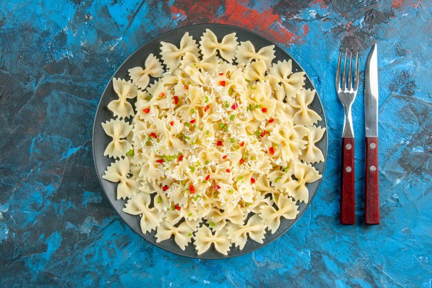 Above view of raw Italian farfalle pasta with vegetables and cutlery set on blue background