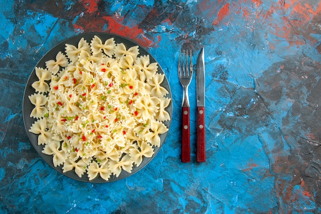 Above view of raw Italian farfalle pasta with vegetables and cutlery set on the right side on blue background