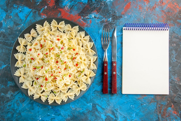 Above view of raw Italian farfalle pasta with vegetables and next to spiral notebook on blue background