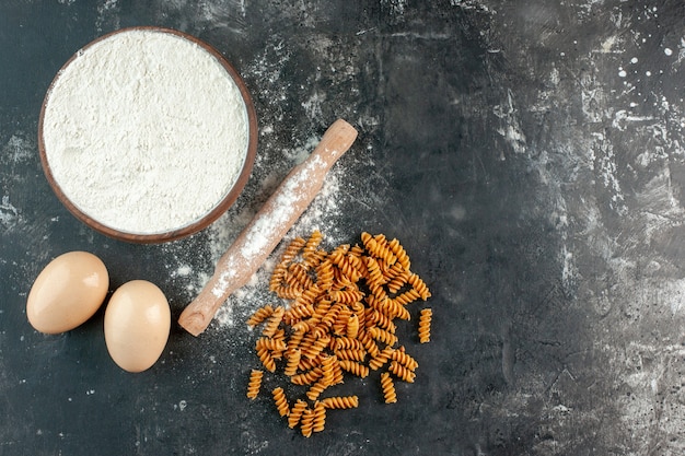 Above view of raw Italian pastas two eggs and flour in a brown bowl rolling-pin on gray background