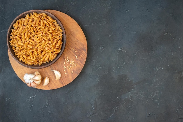 Above view of raw pastas in a brown bowl garlics rice on wooden board on the right side on black background