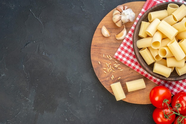 Foto gratuita sopra la vista di pasta cruda all'interno e all'esterno di una ciotola marrone su asciugamano rosso spogliato garlics riso su tavola di legno rotonda pomodori sul lato sinistro su sfondo nero