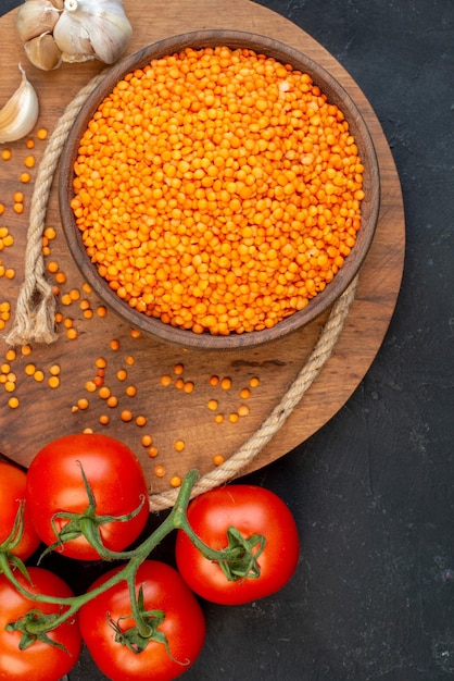 Free photo above view of red lentil in a brown bowl rope garlics on wooden round board and tomatoes with stems on black background