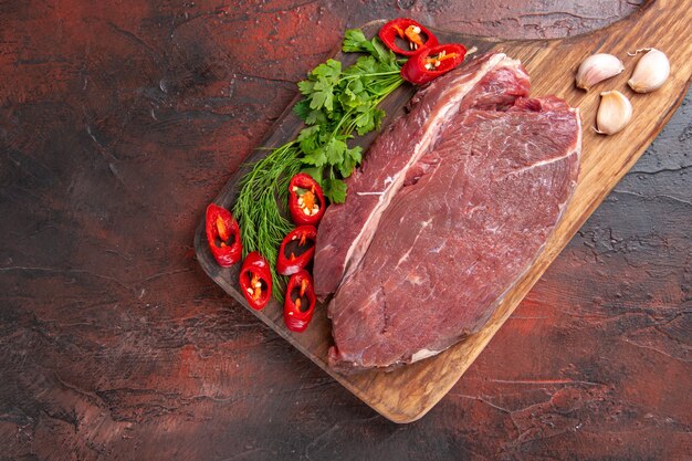 Above view of red meat on wooden cutting board and garlic green chopped pepper on dark background