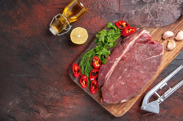 Above view of red meat on wooden cutting board and garlic green chopped pepper fallen oil bottle lemon on dark background