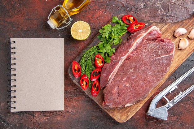 Above view of red meat on wooden cutting board and garlic green chopped pepper fallen oil bottle and notebook on dark background