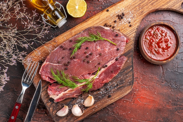 Above view of red meat on wooden cutting board and garlic green fork and knife fallen oil bottle and ketchup on dark background
