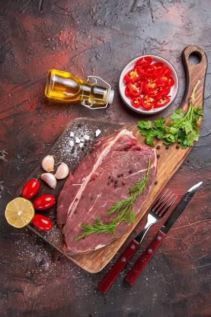 Above view of red meat on wooden cutting board and garlic green lemon chopped pepper tomato fallen oil bottle on dark background