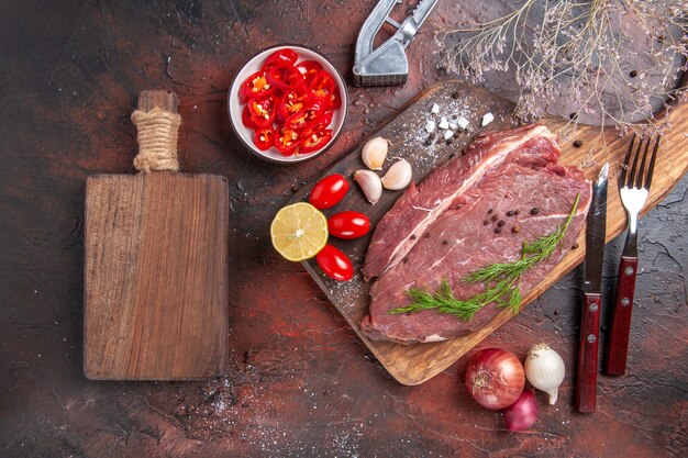 Выше вид красного мяса на деревянной разделочной доске и чеснока, зеленого лимона, лука, вилки и ножа на темном фоне.