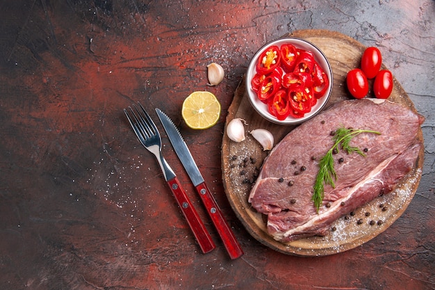 Above view of red meat on wooden tray and garlic green chopped pepper fallen oil bottle fork and knife on dark table