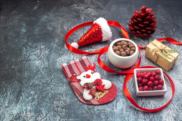 Above view of santa claus hat and cornel chocolate new year sock red conifer cone on the left side on dark surface