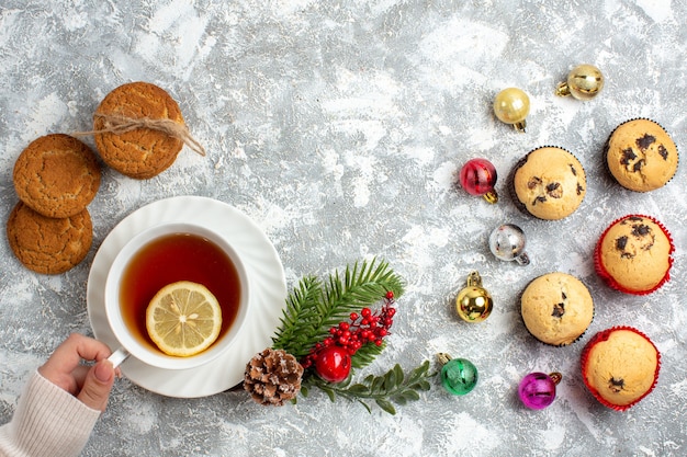 Above view of small cupcakes and decoration accessories fir branches conifer cone hand holding a cup of black tea stacked cakes on ice surface