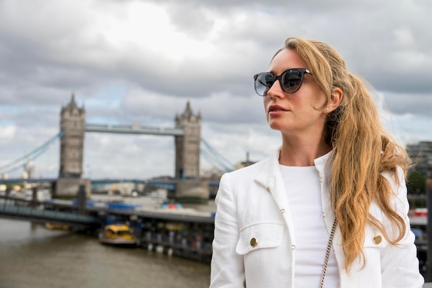 Free photo view of a smiling blonde woman in white jacket and sunglasses with thames and tower bridge on the background united kingdom