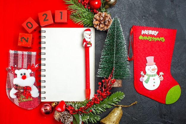 Above view of spiral notebook decoration accessories fir branches xsmas sock numbers on a red napkin and christmas tree on dark background