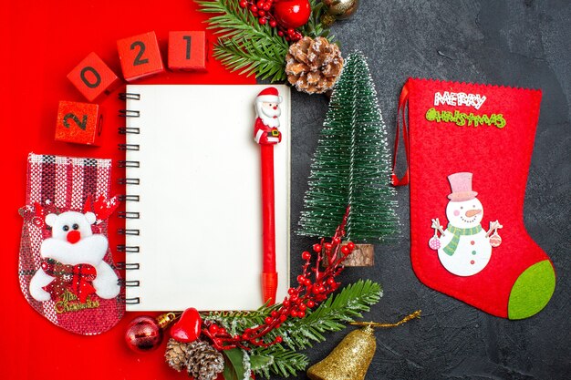 Above view of spiral notebook decoration accessories fir branches xsmas sock numbers on a red napkin and christmas tree on dark background