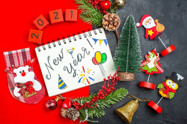 Above view of spiral notebook with new year writing and drawings decoration accessories fir branches xsmas sock numbers on a red napkin and christmas tree on dark background