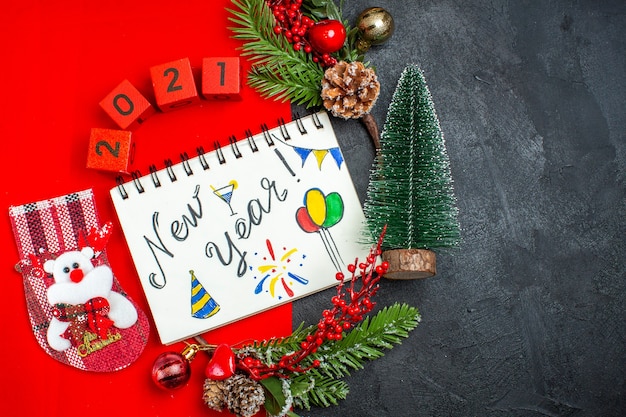 Выше вид спиральной тетради с новогодним письмом и рисунками, украшениями, еловыми ветками, номерами рождественских носков на красной салфетке и рождественской елкой с правой стороны на темном фоне
