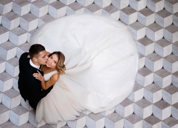 Free photo above view of stylish and serious bridegroom wearing in black suit standing and kissing his wife attractive and cheerful bride