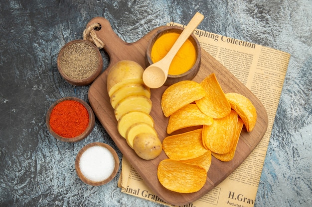 Above view of tasty homemade chips cut potato slices on wooden cutting board and different spices on newspaper on gray table