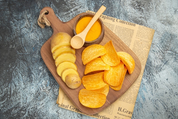 Above view of tasty homemade chips cut potato slices on wooden cutting board on newspaper on gray table