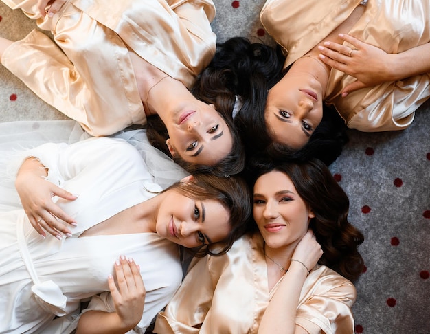 Free photo above view of thoughtful and pretty bride with her friends which wearing in white and beige silk peignoirs lying on floor in room sexually looking and posing at camera during wedding day