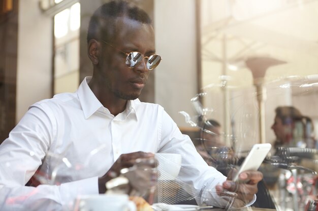 View through window glass of handsome black bisunessman or corporate worker wearing round shades and formal shirt drinking coffee and checking e-mail on mobile phone during break at modern cafe