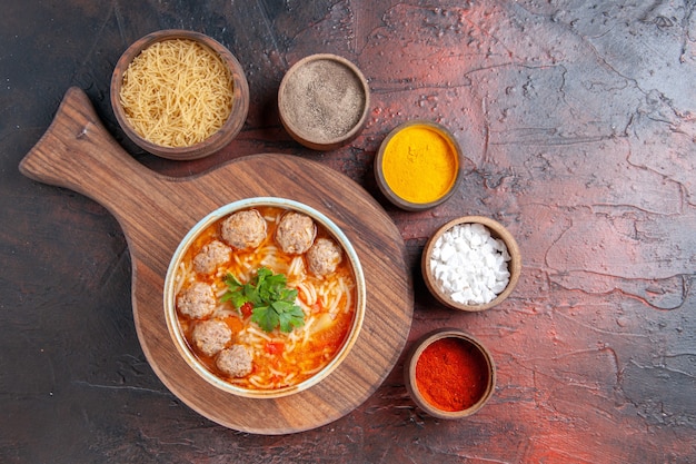 Above view of tomato meatballs soup with noodles in a brown bowl and different spices on dark background