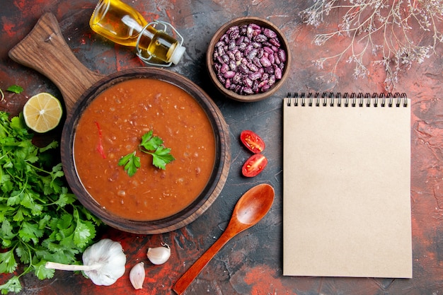 Above view of tomato soup on a cutting board beans oil bottle and notebook on a mixed color table