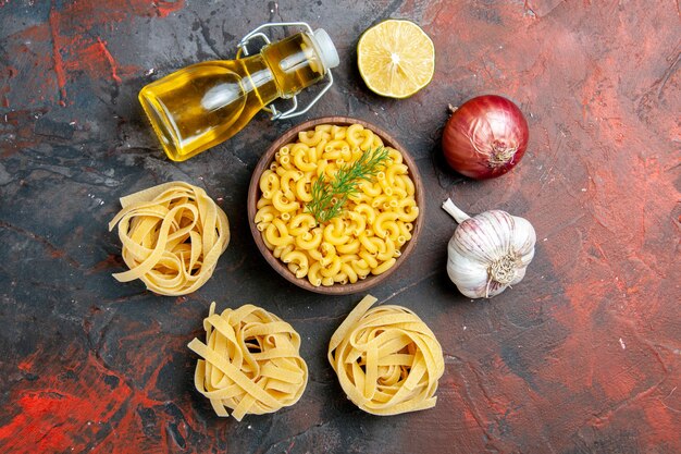 Above view of uncooked three portions of spaghettiand butterfly pastas in a brown bowl and green onion lemon garlic oil bottle on mixed color table