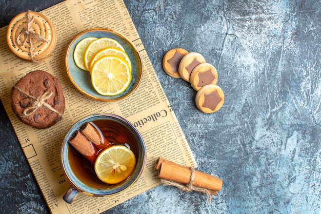 Above view of various cookies and a cup of black tea with cinnamon on an old newspaper on dark background