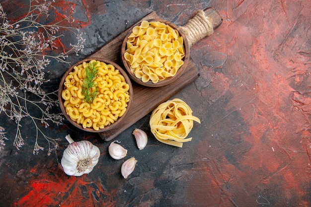 Above view of various types of uncooked pastas on wooden cutting board and garlic on mixed color table