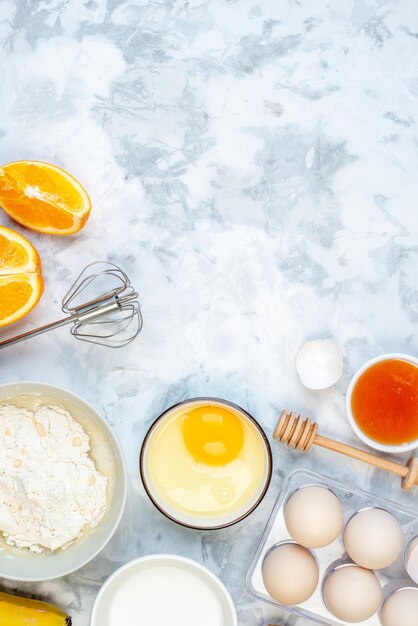 Above view of white flour in a bowl and stainless cooking tool whole cracked eggs fresh oranges on two-toned background