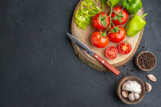 Above view of whole cut chopped green peppers and fresh tomatoes knife on wooden cutting board pepper garlics on the left side on black surface