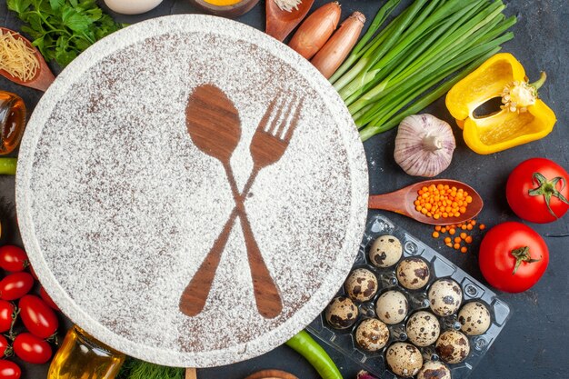 Above view of wooden board with flour drawn fork and knife among organic vegetables eggs fallen oil bottle pepper on black