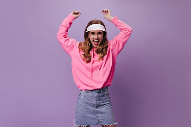 Vigorous woman in pink hoodie and cap shouts on purple background Emotional girl in denim skirt dancing on isolated backdrop