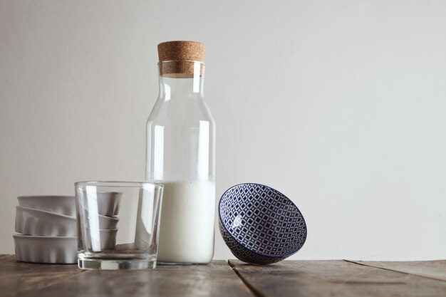 Vintage bottle closed cork with milk on aged wooden table near whiskey rox transparent glass, white ceramic plates and bowl with glazed pattern, isolated on white