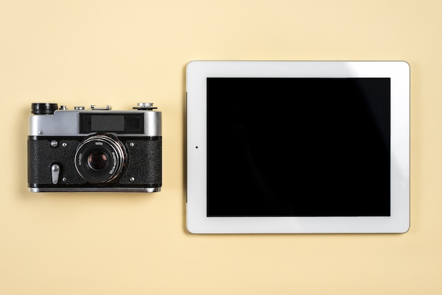 Vintage camera near the digital tablet with blank black screen display on beige background