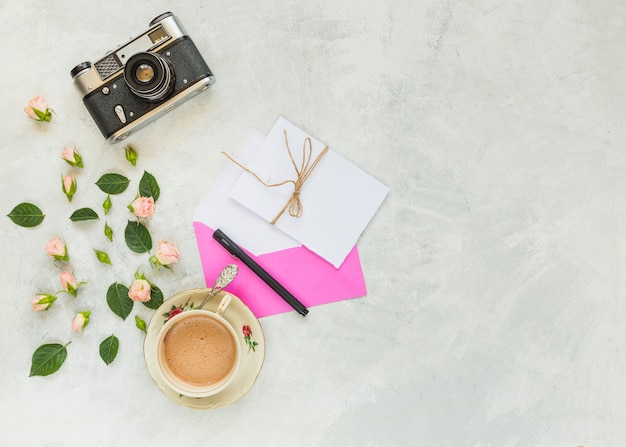 Винтажная камера; розовая роза; зеленые листья; конверт; бумага; ручка и чашка кофе на бетонном фоне